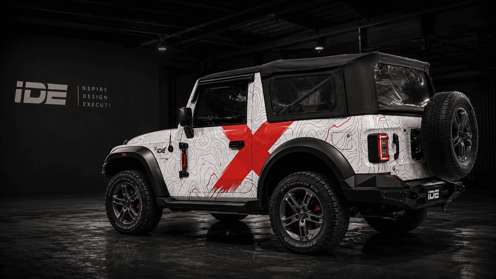 An off-road vehicle finished in a white topographic-line graphic with a bold red 'X', photographed in a dark studio beside the IDE logo